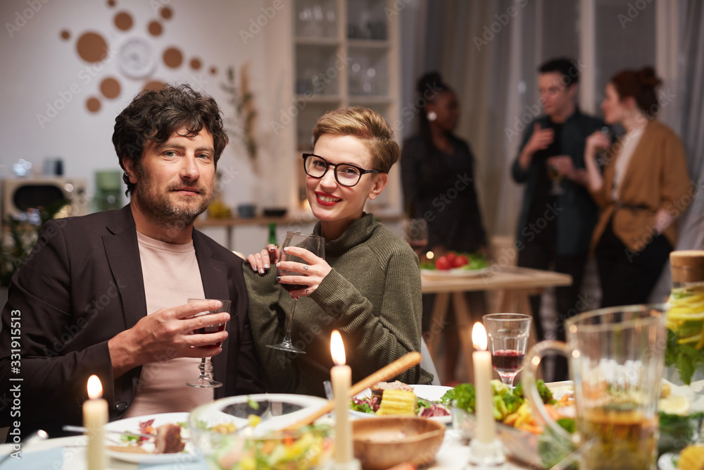 Portrait of young couple smiling at camera while sitting at the table and drinking wine during the party Portrait of young couple smiling at camera while sitting at the table and drinking wine during the party