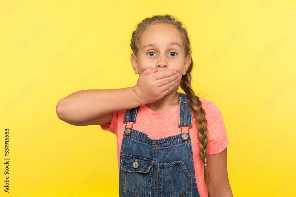 I won't tell! Portrait of scared little girl with braid in denim overalls covering mouth with hand, afraid to say secret, child terrified to speak. indoor studio shot isolated on yellow background I won't tell! Portrait of scared little girl with braid in denim overalls covering mouth with hand, afraid to say secret, child terrified to speak. indoor studio shot isolated on yellow background