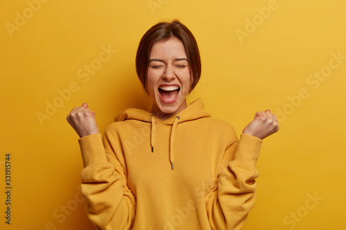 Lucky positive woman feels happy and relieved, achieves reward, clenches fists with triumph, celebrates great amazing news, exclaims with joy, wears casual hoodie, isolated on yellow background photo