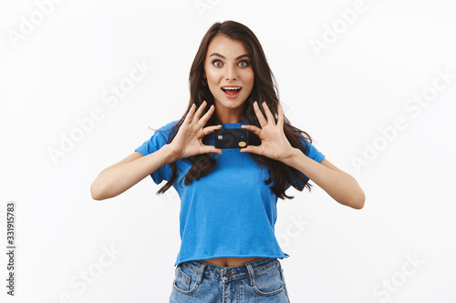 Excited smiling and pumped brunette female in basic blue t-shirt, holding black credit card with both hands, inviting friend shopping together, got money, paycheck, standing white background