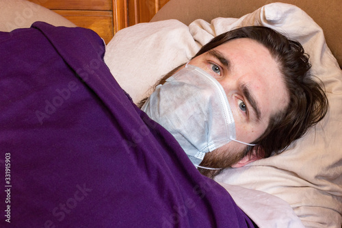 a man with dark hair and a disposable mask to protect against the virus, lies at home in bed, isolating himself from other people. fever, headache and nausea are symptoms - take care of your health