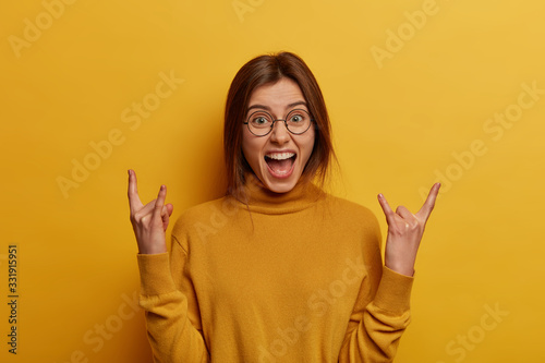 Lively charismatic upbeat woman raises hands and makes rock n roll gesture, shares positive vibes, attends concert of favorite band, goes crazy partying, dressed casually, isolated on yellow wall