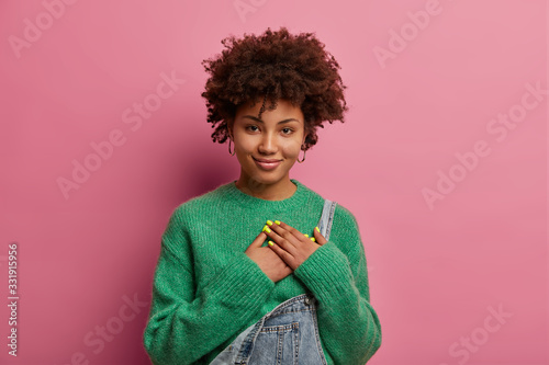 Touched thankful ethnic woman keeps hands on heart, expresses gratitude and kindness for nice gift, smiles gently, wears green sweater and overalls, thanks for help, poses against pink background. photo