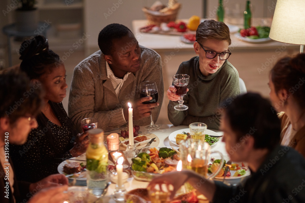 Multiethnic group of young people sitting at the table drinking wine eat holiday dinner and talking to each other