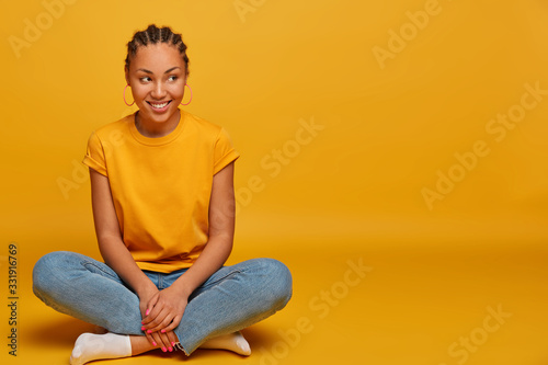 Photo of attractive carfree Afro American woman sits crossed legs, happy to hear interesting stories, takes part in conversation, poses against yellow background, copy space for your advertisement © wayhome.studio 
