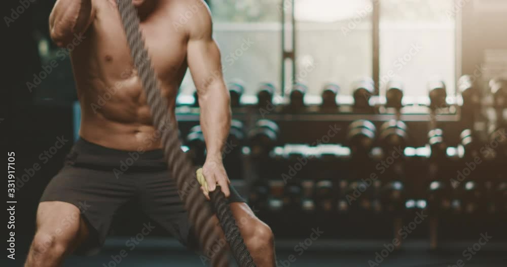 Attractive muscular man working out in the gym using battle ropes with high intensity, determined fitness man achieving his goals