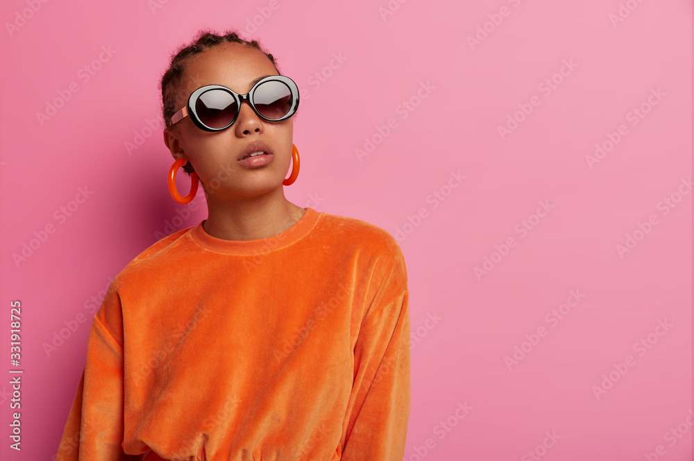Horizontal shot of dark skinned female model wears stylish velvet orange sweater and earrings, big sunglasses, poses serious, buys clothes of last trends, poses against pink background, empty space Horizontal shot of dark skinned female model wears stylish velvet orange sweater and earrings, big sunglasses, poses serious, buys clothes of last trends, poses against pink background, empty space