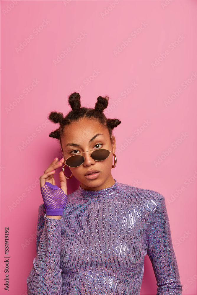 Self confident attractive female has funny hairstyle with buns, keeps hands on shades, wears glittering jumper, looks directly at camera, poses against pink pastel wall, empty space. People, style Self confident attractive female has funny hairstyle with buns, keeps hands on shades, wears glittering jumper, looks directly at camera, poses against pink pastel wall, empty space. People, style