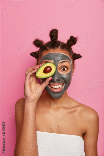 Taking care of your skin in natural way. Positive young woman applies clay mask on face  holds piece of avocado in front of eye  smiles happily  enjoys cosmetic treatment  cares about complexion