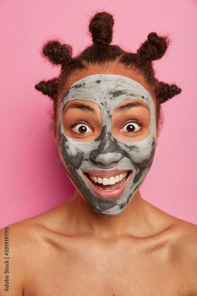 Close up portrait of shirtless positive woman has happy surprised reaction, beaming smile, perfect white teeth, wears clay mask for smooth face skin, isolated on pink background. Wellness concept