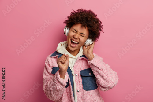 Photo of joyful Afro American woman sings song loudly, enjoys awesome music in stereo headphones, dressed in casual hoodie and jacket, keeps hand as microphone near mouth, isolated on pink wall