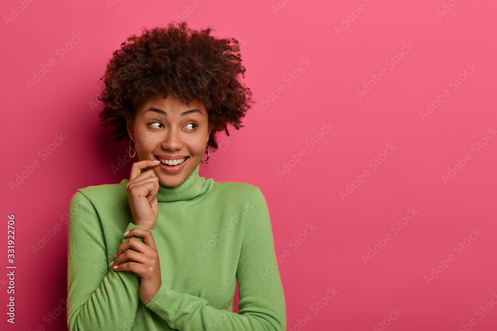 Studio shot of cheerful pleased woman looks aside with tender smile, keeps finger near mouth, wears casual green turtleneck, happy to see something into distance, isolated on pink background Studio shot of cheerful pleased woman looks aside with tender smile, keeps finger near mouth, wears casual green turtleneck, happy to see something into distance, isolated on pink background