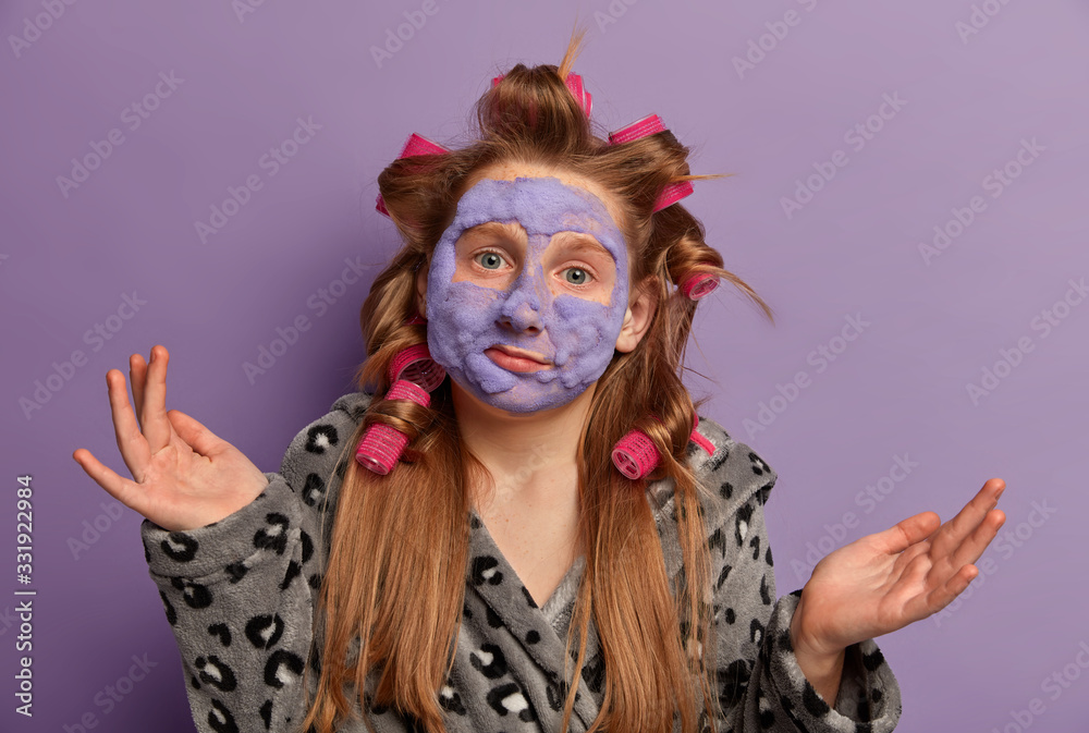 Photo of confused ginger preteen girl spreads palms, feels hesitant, wears curlers for making hairstyle, applies beauty mask, dressed in domestic clothes, doesnt know how to improve skin condition