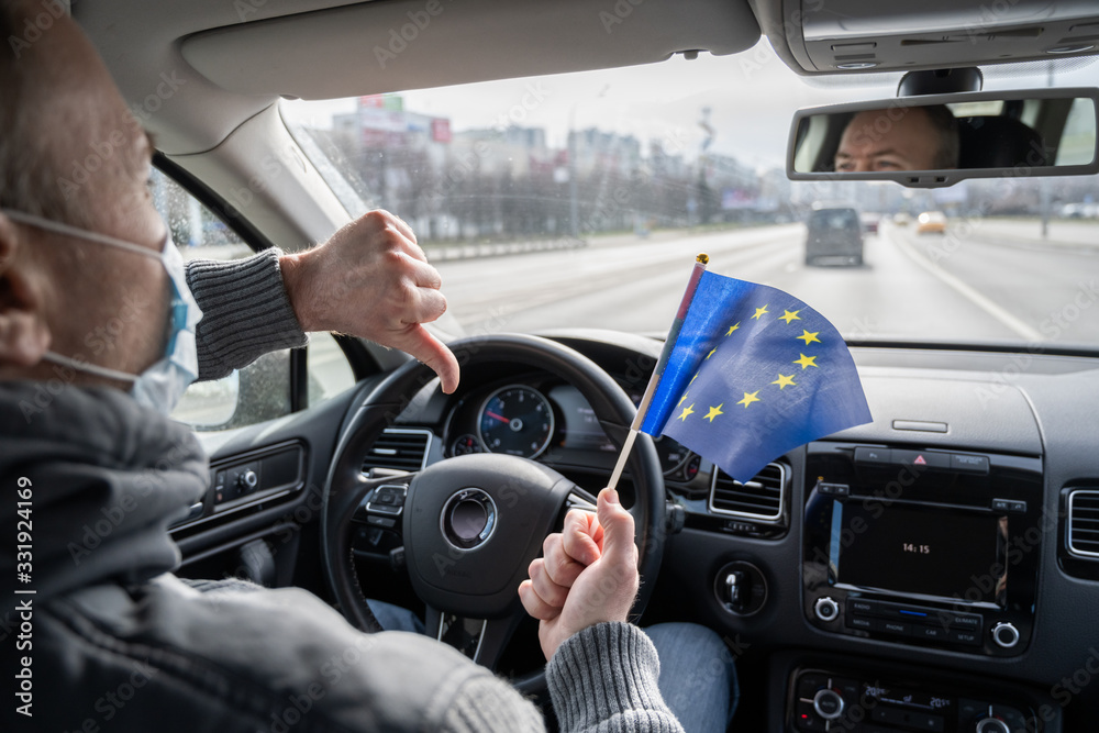 Middle aged man in the medical mask showing thumbs down with European Union (EU) flag driving a car. symbol of adverse epidemiological situation in countries. Coronavirus. Pandemic