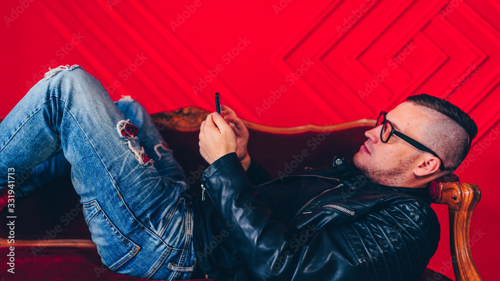 Handsome young man with glasses resting on small sofa. Adult charming male flipping through various news in mobile phone, lying on armchair.