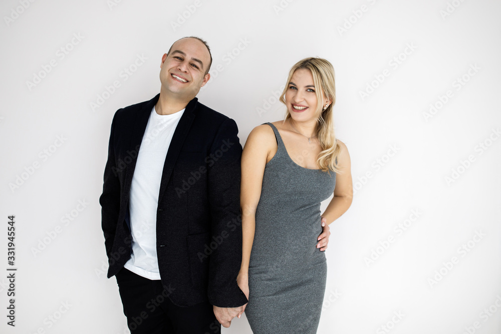 Young pretty blonde, expectant mother, stands next to her husband. Young couple, man and woman, beautifully and elegantly dressed, standing in studio on a white background. Pragnancy concept. Young pretty blonde, expectant mother, stands next to her husband. Young couple, man and woman, beautifully and elegantly dressed, standing in studio on a white background. Pragnancy concept.