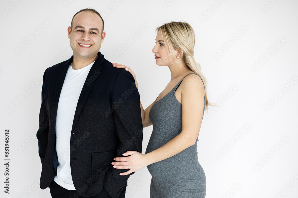 Young pretty blonde, expectant mother, stands next to her husband. Young couple, man and woman, beautifully and elegantly dressed, standing in studio on a white background. Pragnancy concept. Young pretty blonde, expectant mother, stands next to her husband. Young couple, man and woman, beautifully and elegantly dressed, standing in studio on a white background. Pragnancy concept.