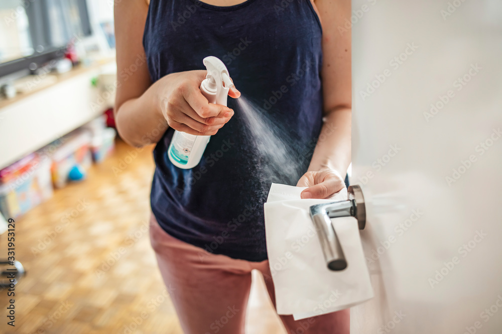 Woman cleaning a door handle with a disinfection spray and disposable wipe. Woman sanitizing door handle with antibacterial spray. Girl staying at home during coronavirus outbreak - obrazy, fototapety, plakaty 