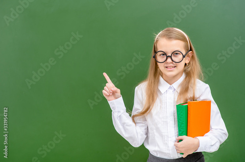 Smart girl holds books and points away on empty green chalkboard. Empty space for text © Ermolaev Alexandr