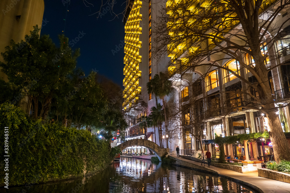 Naklejka premium The popular riverwalk in a night scene in downtown San Antonio, Texas, with an arch bridge and tall buildings along the river reflected in the water.