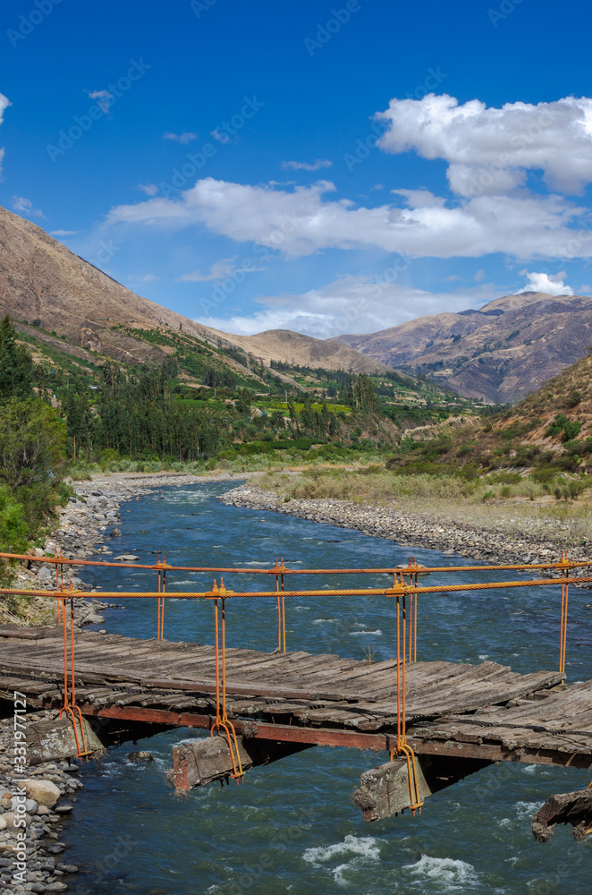 wooden suspension bridge held by ropes over river in valley surrounded by mountains.