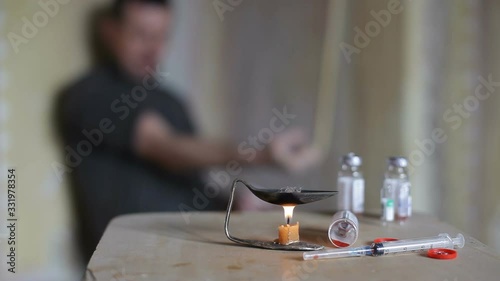 An addict injects a drug. In the foreground, a candle under a spoon, a bottle of drugs and a syringe are lit