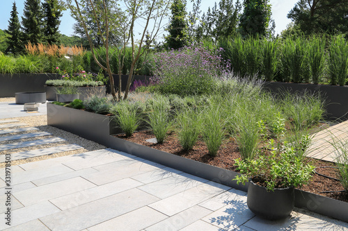 Moderne Garten- und Terrassengestaltung im Materialmix:Terrassen aus Holz, Steinplatten und Schotter sowie Metall Pflanzgefäße mit Gräsern, Bäumen und blühenden Kübel Pflanzen	