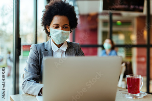 Black businesswoman wearing protective mask on her face while working on laptop in a cafe, photo