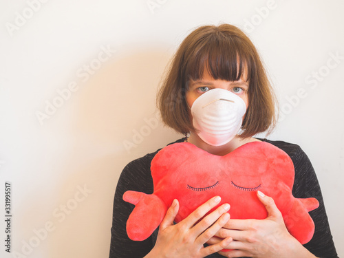 Frustrated caucasian young woman wearing face mask and hugging heart shaped plush toy as symbol of support and hope staying home during Coronavirus pandemic quarantine photo