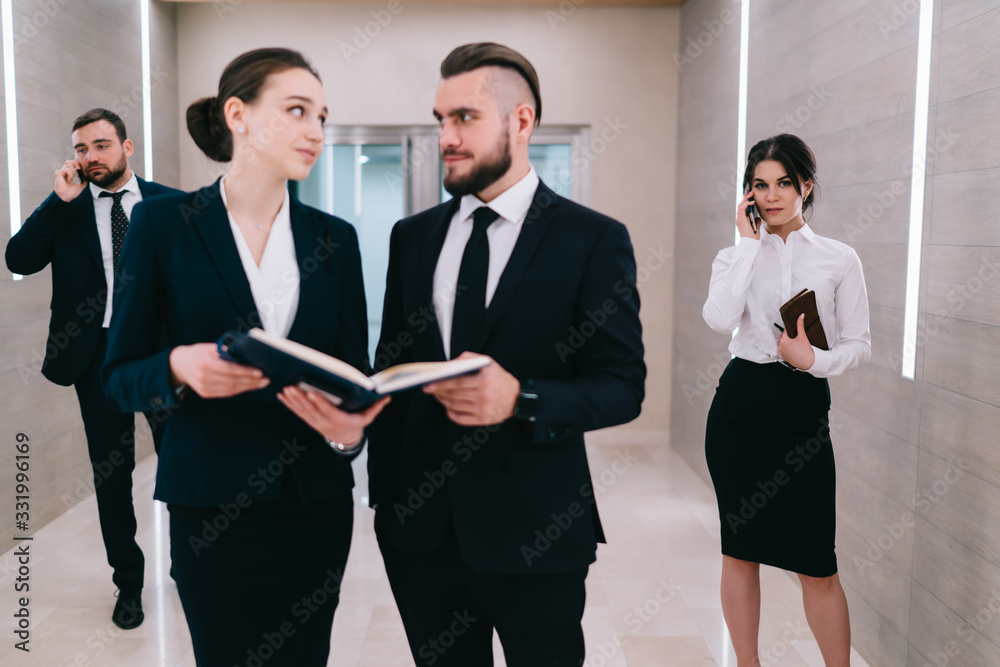 Doubtful coworkers discussing document and using smartphone while standing in office lobby Doubtful coworkers discussing document and using smartphone while standing in office lobby