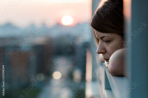 Young sad woman looking outside through balcony of an apartment building