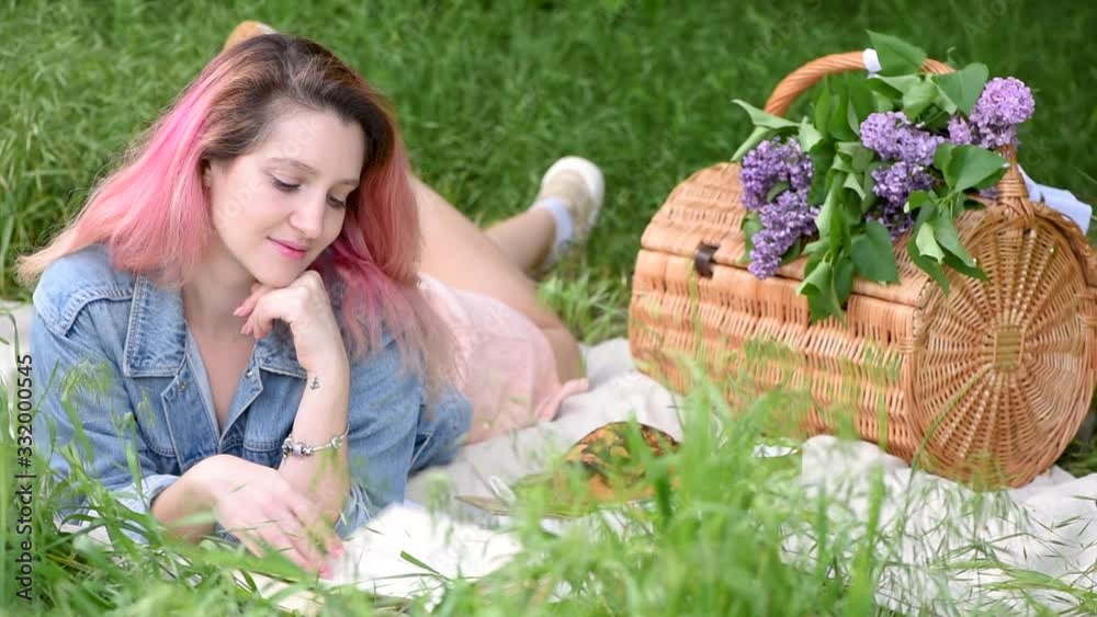 Pink-haired attractive girl in a jeans jacket and a linen dress is sitting on the ground on a picnic with a bouquet of lilac and a book Pink-haired attractive girl in a jeans jacket and a linen dress is sitting on the ground on a picnic with a bouquet of lilac and a book