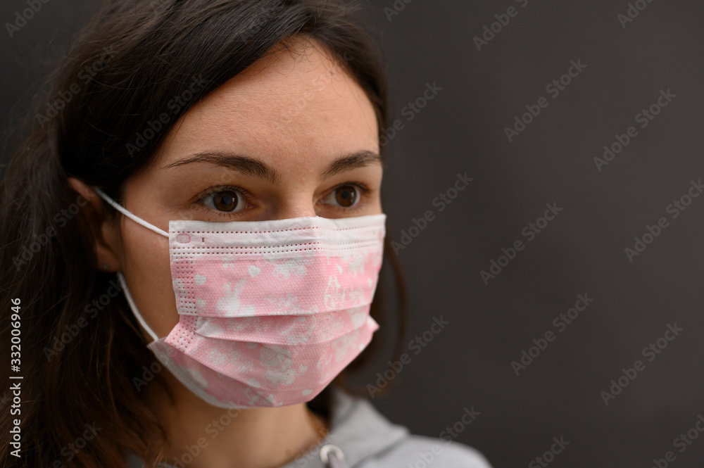 Brunette girl in a disposable medical mask on a dark background. A piercing glance to the side. Close-up. Horizontal view. Brunette girl in a disposable medical mask on a dark background. A piercing glance to the side. Close-up. Horizontal view.