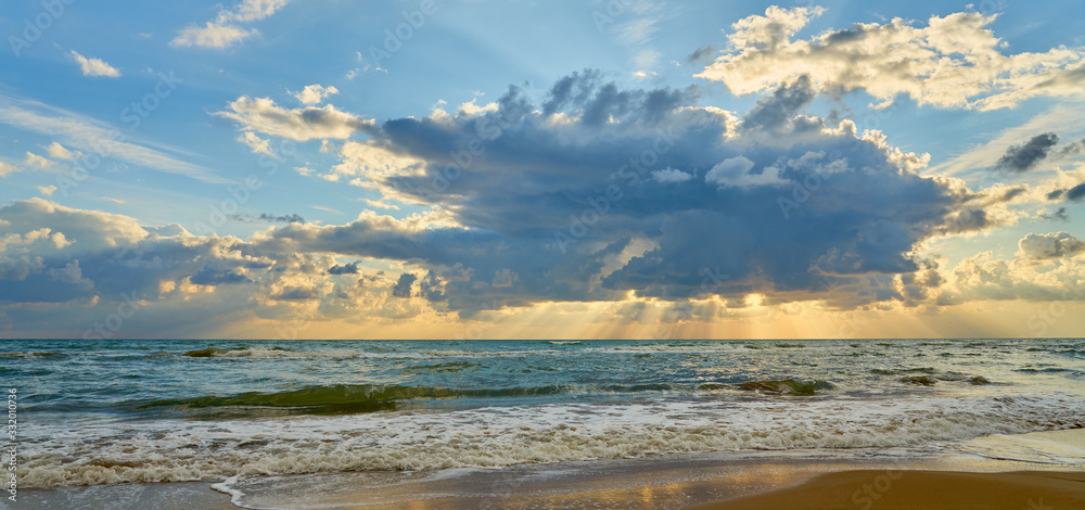 Colorful sunset at the tropical sandy beach, sun behind the clouds and waves with foam hitting sand.