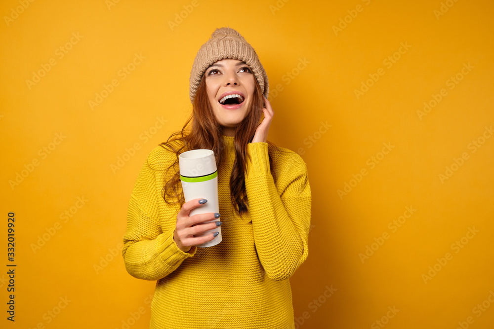 A beautiful brunette in a yellow sweater and hat stands on a yellow background with a white thermos in hands and looks up joyfully A beautiful brunette in a yellow sweater and hat stands on a yellow background with a white thermos in hands and looks up joyfully