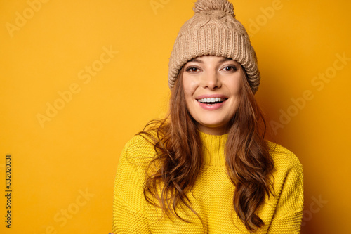 A beautiful brunette with clean skin in a yellow sweater and hat stands on a yellow background smiling joyfully.