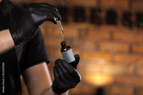 Man holding essential oil for beard or aromatherapy in glass bottle on hand and dripping oil. man with black gloves in barber shop. copy space on bottle photo