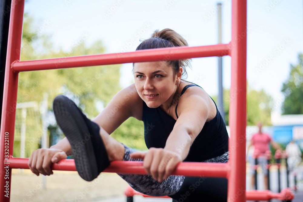 Obraz premium Young brunette woman, wearing black fitness overall, doing stretching exercises at swedish wall by city lake in summer. Fit sportswoman on outside training. Healthy energetic lifestyle concept.