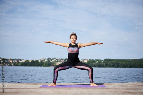 Young brunette woman with bare feet  wearing black and purple fitness outfit  stretching on violet yoga mat outside on wooden pier in summer. Fit girl  doing yoga poses by lake. Healthy lifestyle.