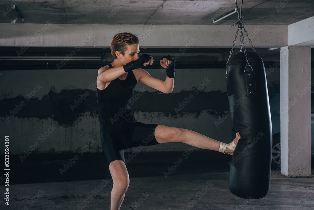 Caucasian woman in sportswear and with black bandages kicking the boxing bag inside a garage