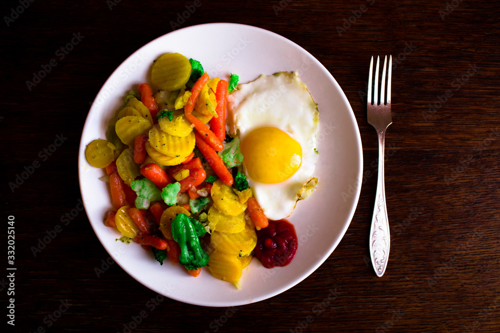 Fried egg with delicious vegetables on a white plate and a metal fork located on a dark wooden textured table. Fried egg with delicious vegetables on a white plate and a metal fork located on a dark wooden textured table.