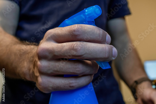 A tan hairy arm person holds up a blue spray bottle filled with alcohol to protect against germs and bacteria that may infest the objects around them.
