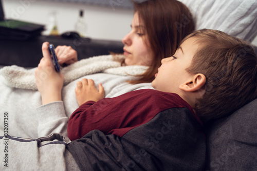 Side view on caucasian boy little kid mother and son lying on the sofa or bed playing games on mobile smart phone or making video call typing sms texting messages at home in evening during quarantine photo