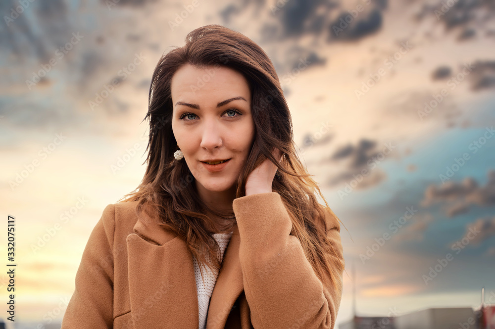 Portrait of a brunette girl standing in a park in a coat walking a street on a background of spring sky