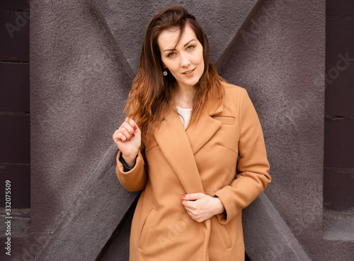 Portrait of a brunette girl standing in spring in a beige coat walk a street on a background of a dark purple wall of a building