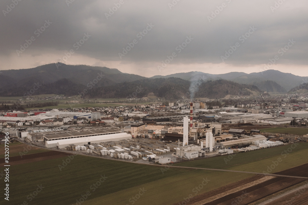 Aerial photo of a factory sitting between the green fields. Visible smoke coming out from a smokestack. Factory in the nature on a dull day, concept of dirty factory in nature.
