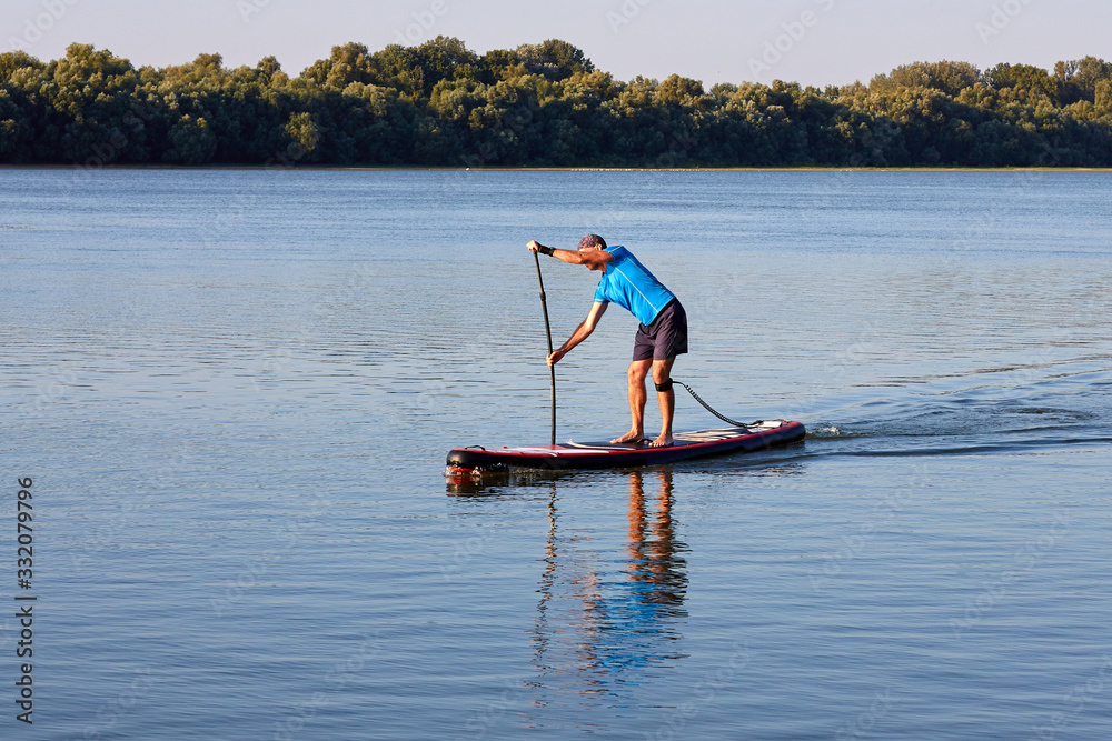 Naklejka premium Joyful man is training on a SUP board on Danube river during sunny morning. Stand up paddle boarding - awesome active recreation in nature