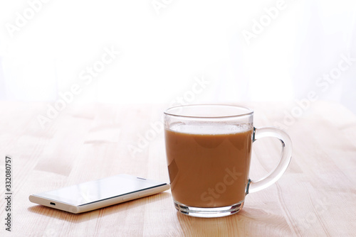 glass coffee with milk mug and smartphone lit by morning sunlight on a wooden table, copy space