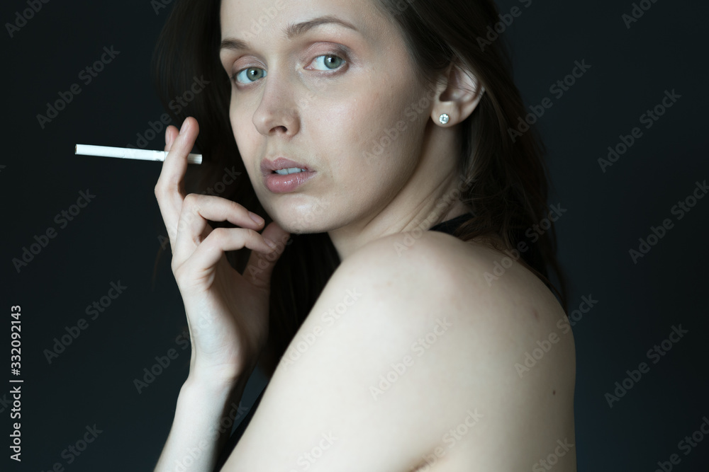 Fototapeta premium Portrait of a young woman with long hair, who looks directly into the camera, holding a cigarette near her mouth, on a gray background