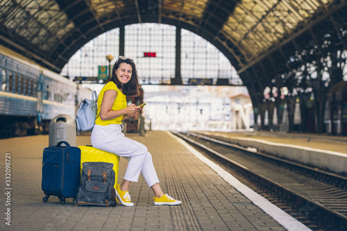 smiling pretty woman sitting on yellow suitcase with wheels at railway station